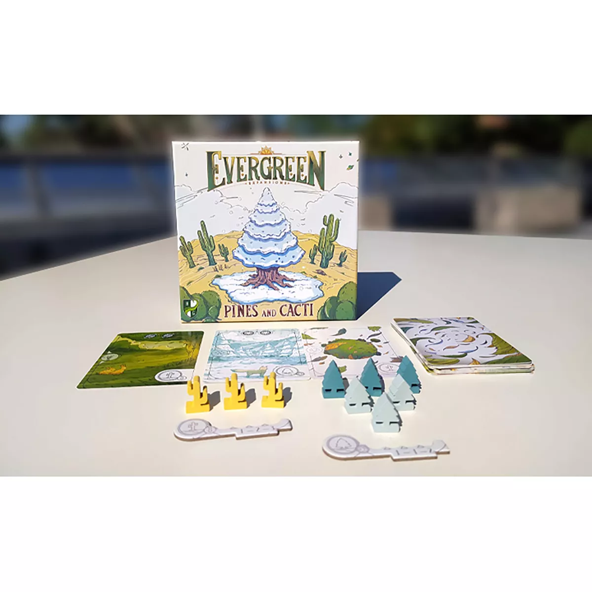 The Evergreen Pines and Cacti Expansion board game box stands upright with various game components including cards, yellow cacti, and blue-green tree tokens spread on a white table