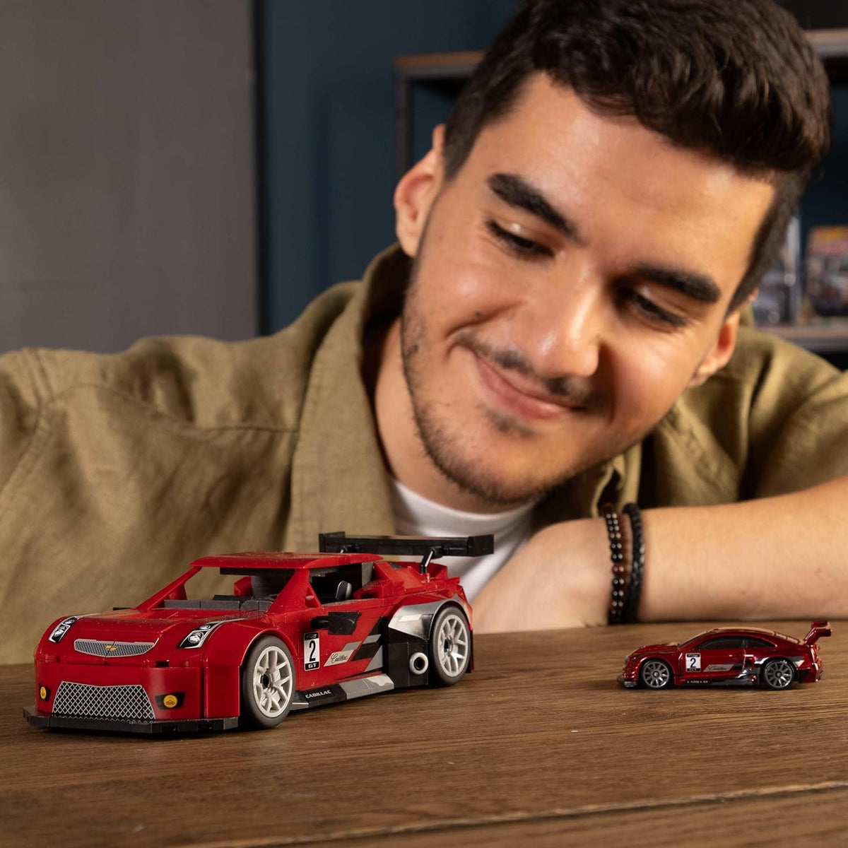 Smiling man looks at a large red LEGO Cadillac race car model next to a smaller die-cast version on a table.