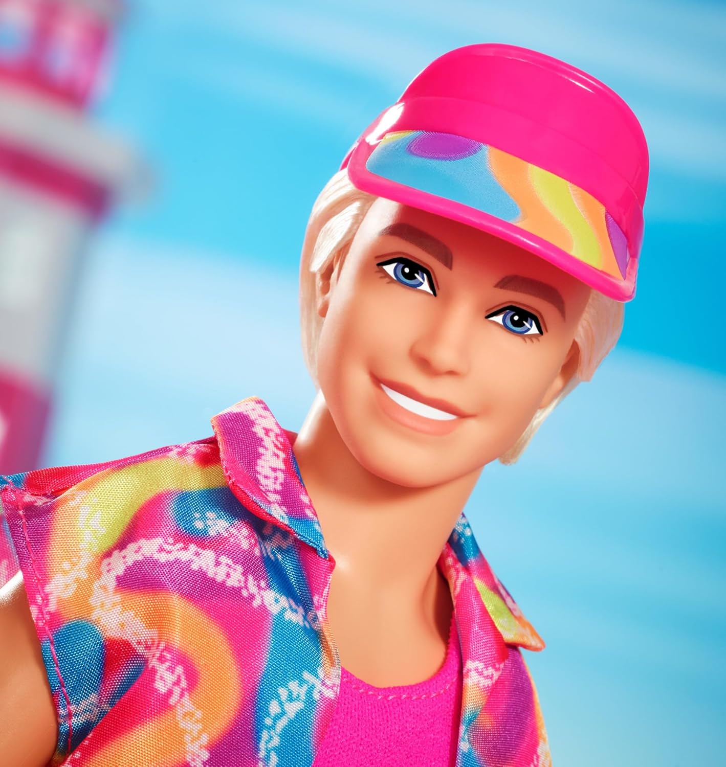 Close-up of a smiling Ken doll in a pink visor and colorful tie-dye shirt -  with a blurry lighthouse in the background