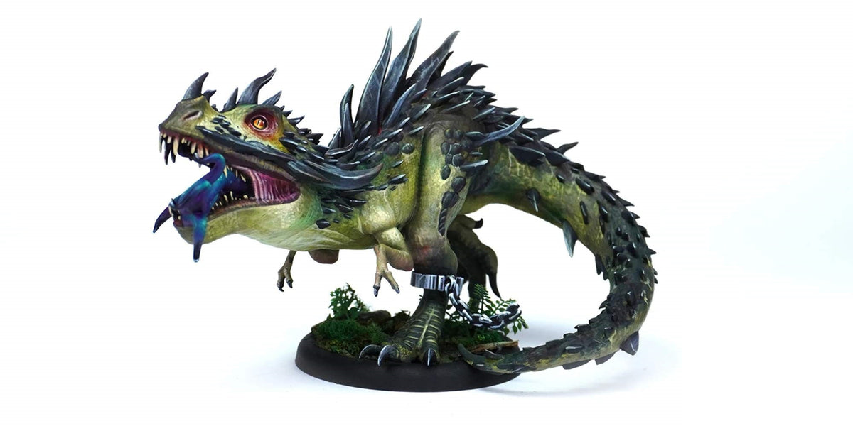 A green, spiky Malifaux Explorer&#39;s Society Malisaurus Rex miniature devouring a blue bird, chained at the leg, standing on a mossy base