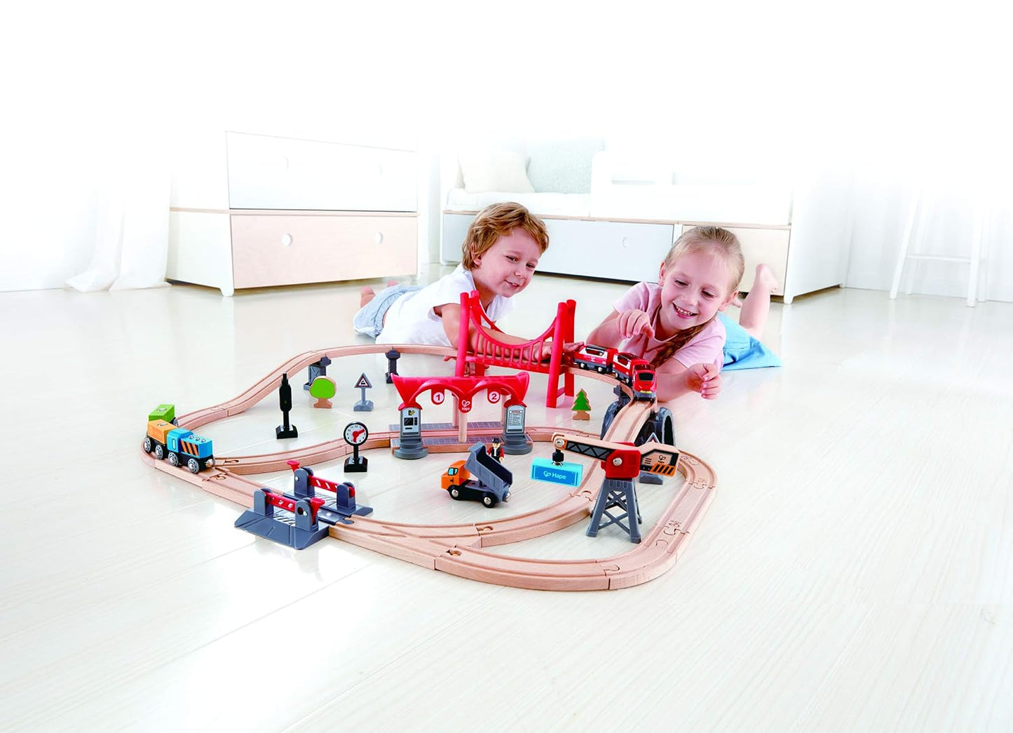 Two smiling children lie on the floor playing with a large wooden train set and red bridge