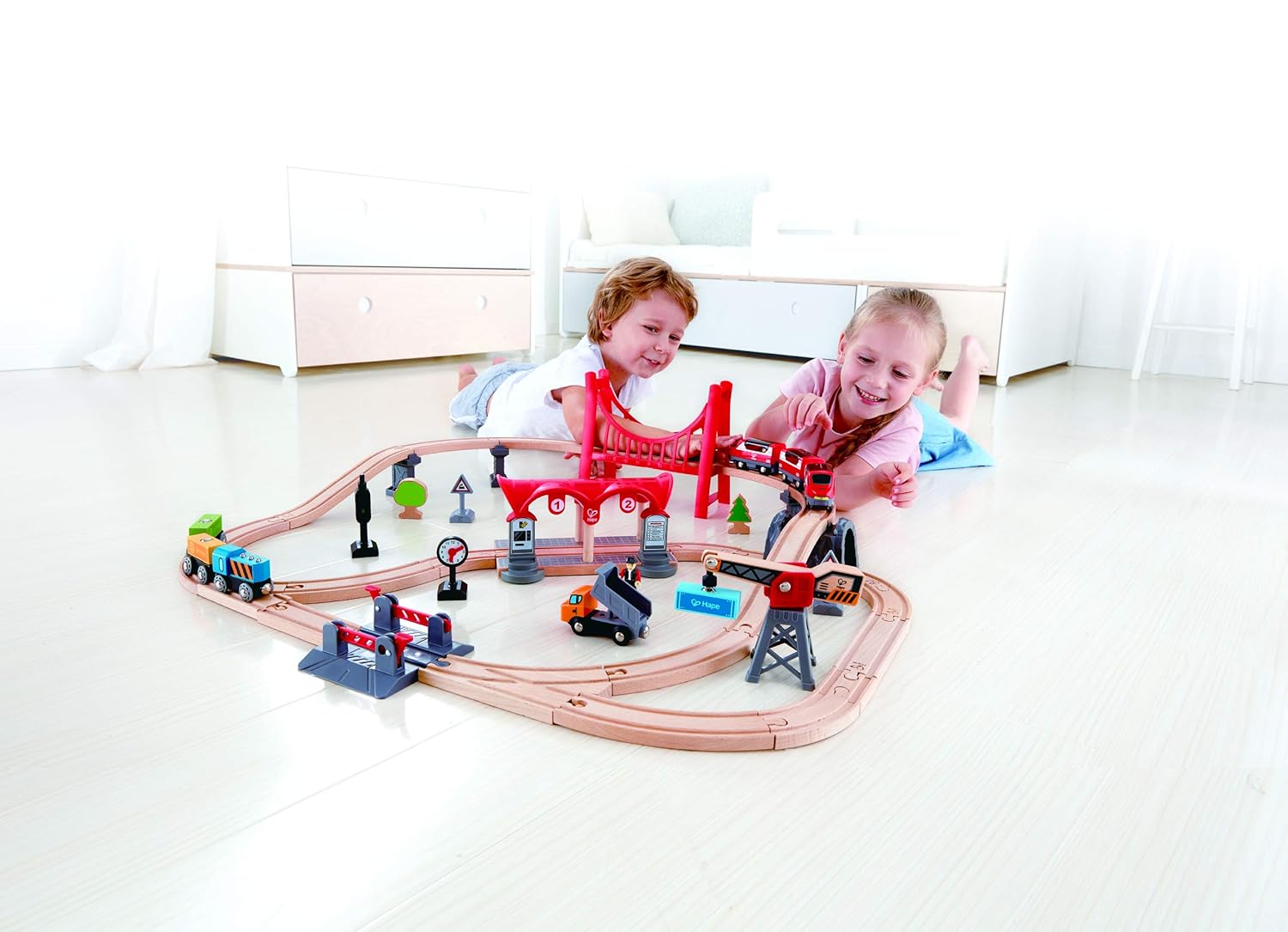 Two smiling children lie on the floor playing with a large wooden train set and red bridge