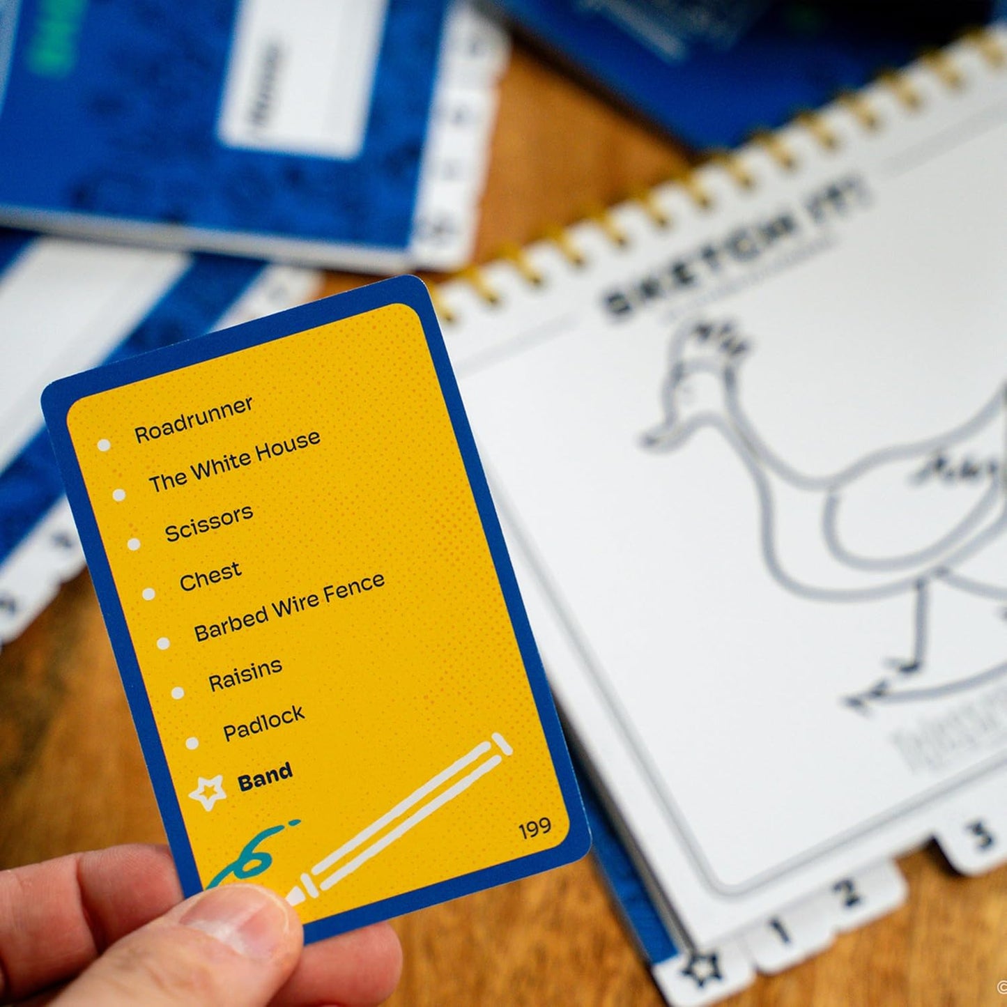Hand holding a yellow game card (listing words) over a sketchpad with a drawing of a goose/bird