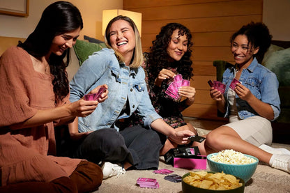 Four laughing women playing a card game together in a cozy living room setting with snacks
