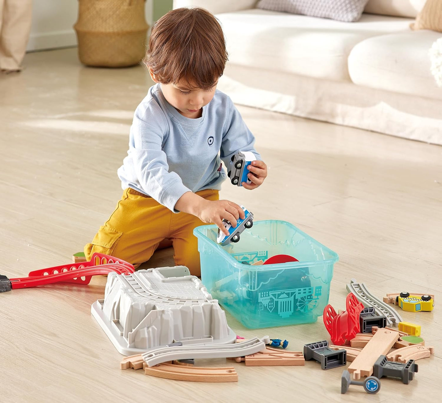 Toddler playing with wooden train tracks and toy cars on the floor, placing a car in a storage bin