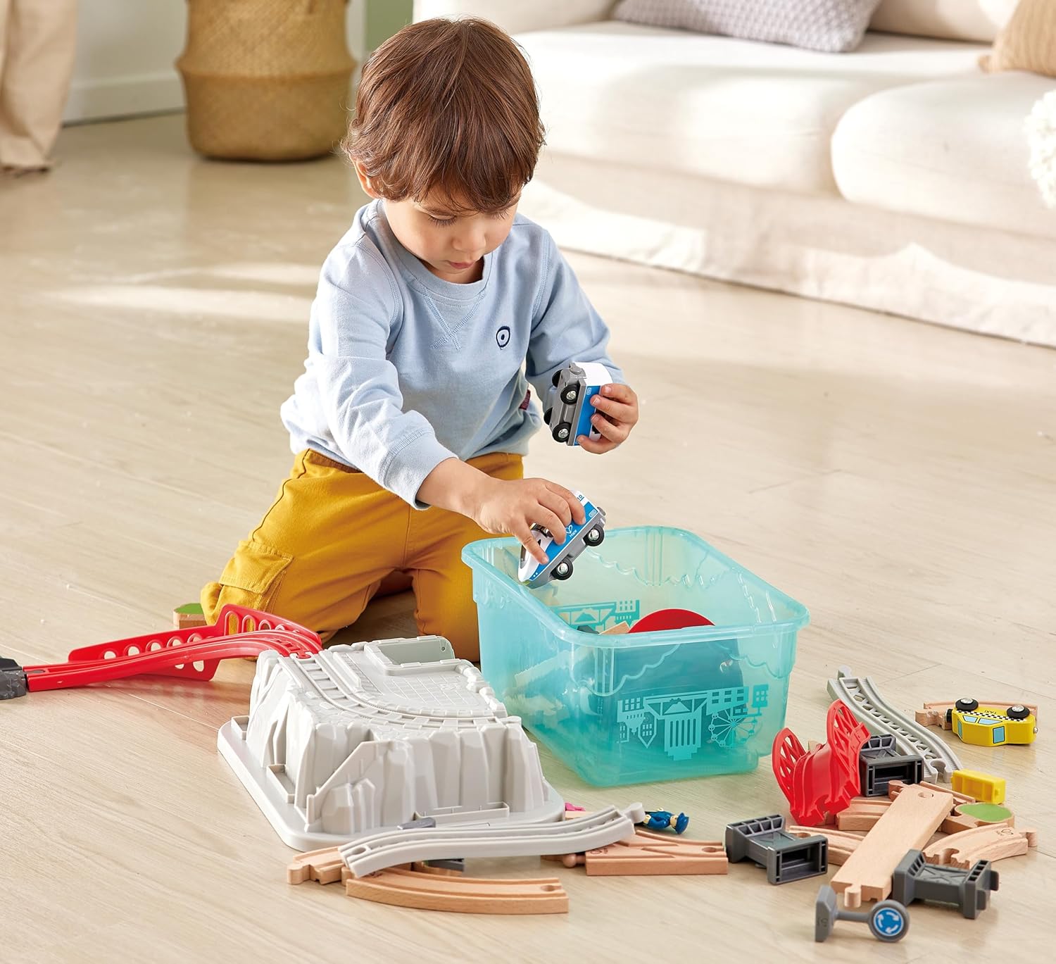 Toddler playing with wooden train tracks and toy cars on the floor, placing a car in a storage bin