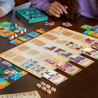 Two players interact with components of a tile-laying board game, featuring colorful cards, tiles, and wooden tokens