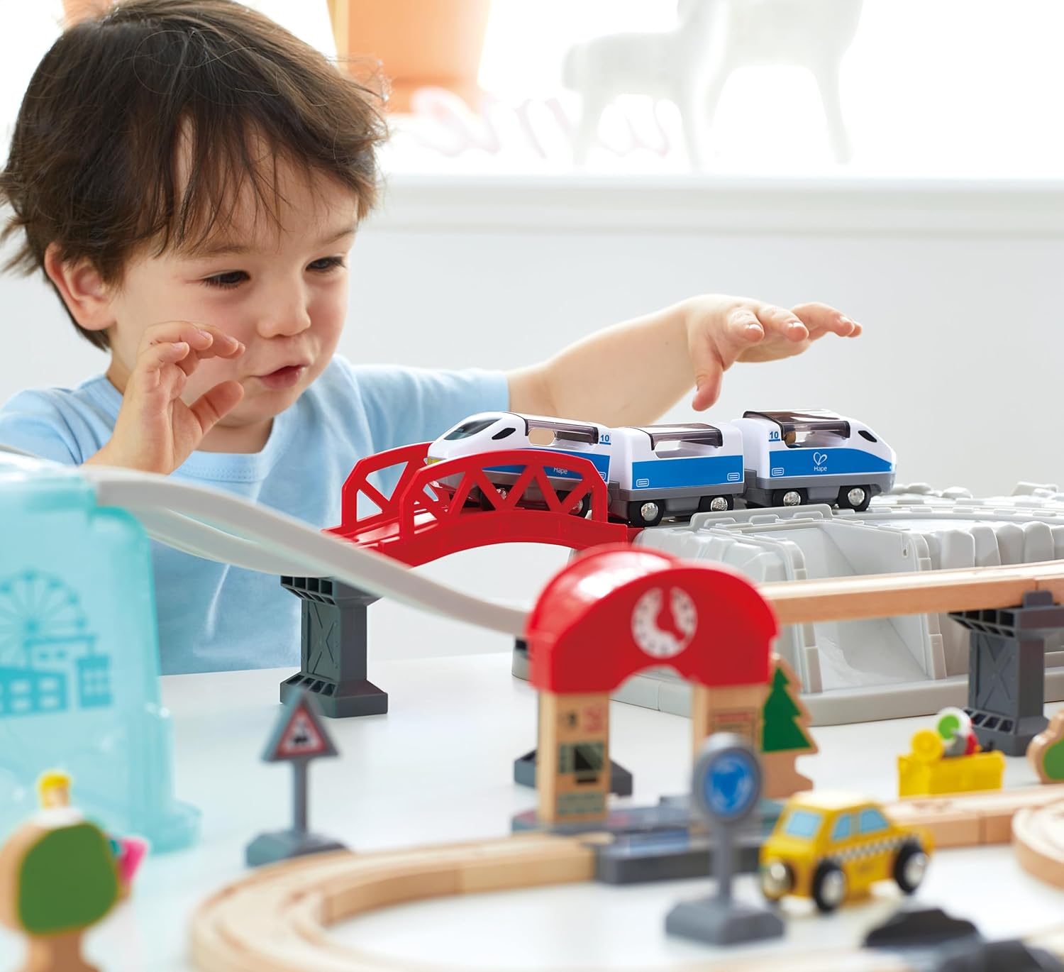 Young child playing with a toy electric train crossing a red bridge on a wooden track set