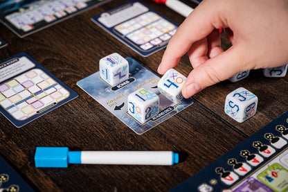 Player's hand selecting a custom die next to game cards and components on a dark wooden table