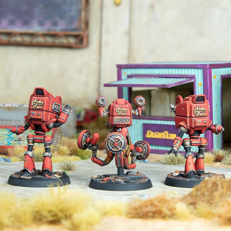 Three red Nuka Cola robot miniatures: two bipedal, one with circular arms. A "Quantum" branded kiosk sits in a dusty wasteland background