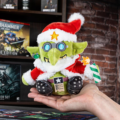Santa Grot plush with goggles and a lit firecracker, held above Warhammer games