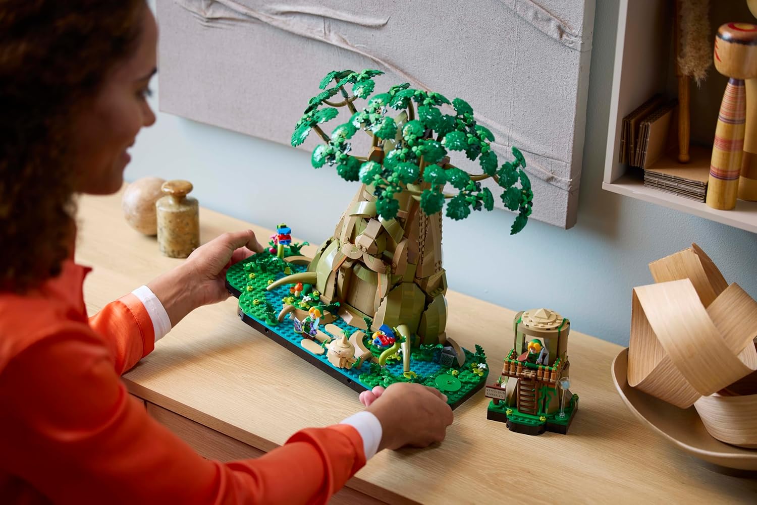 Woman admires a detailed, green LEGO diorama of a large fantasy tree on a wooden desk