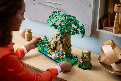 Woman admires a detailed, green LEGO diorama of a large fantasy tree on a wooden desk
