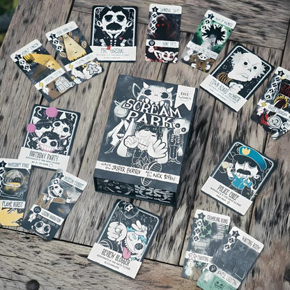 A detailed overhead shot of the Scream Park board game box and numerous brightly illustrated black and white character and action cards scattered on a rough wood surface