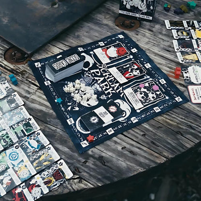 Scream Park board game components including the black cloth playmat, stacked scene cards, and white skull-like tokens are spread across a distressed wooden surface