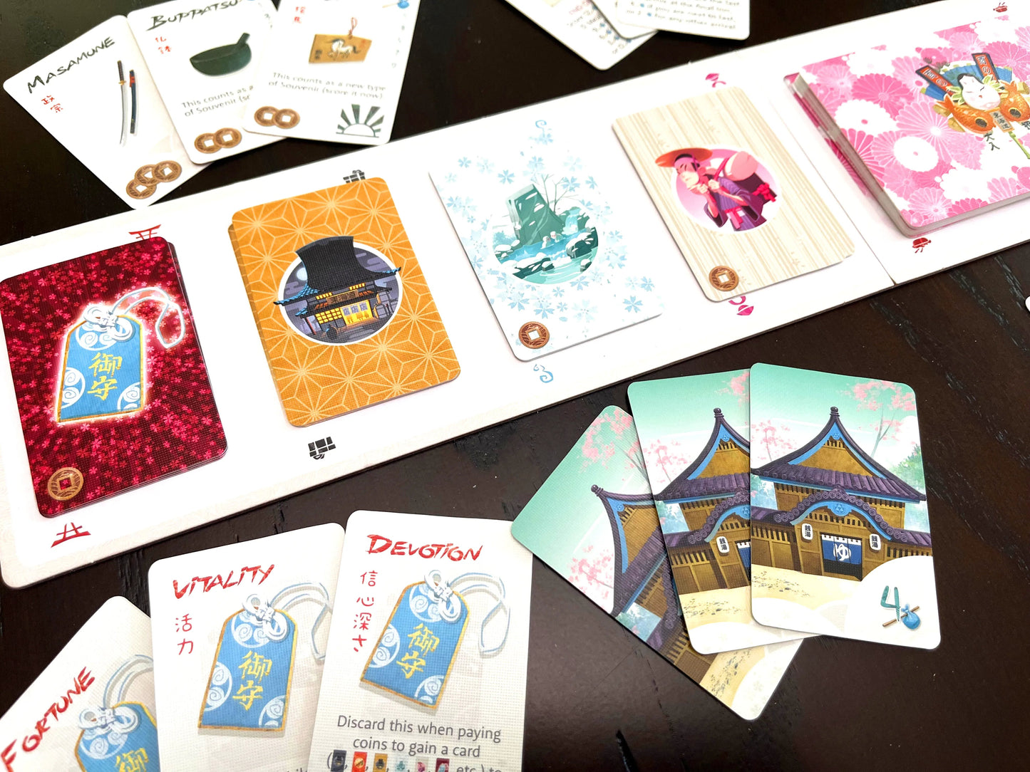 Close-up of Tokaido board game components displaying Crossroads Omamori cards like Devotion and Vitality next to Matsuri temple artwork and souvenir tokens