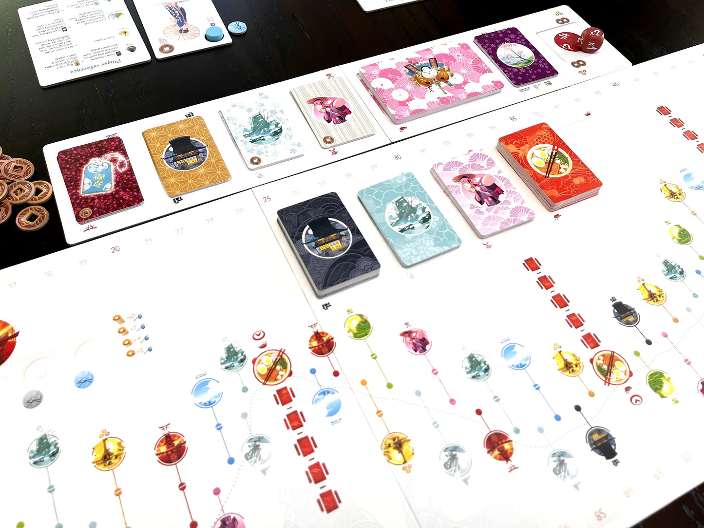Components of the Tokaido game, including the numbered white board track, colorful souvenir and encounter card stacks featuring Japanese imagery, and scattered tokens and dice