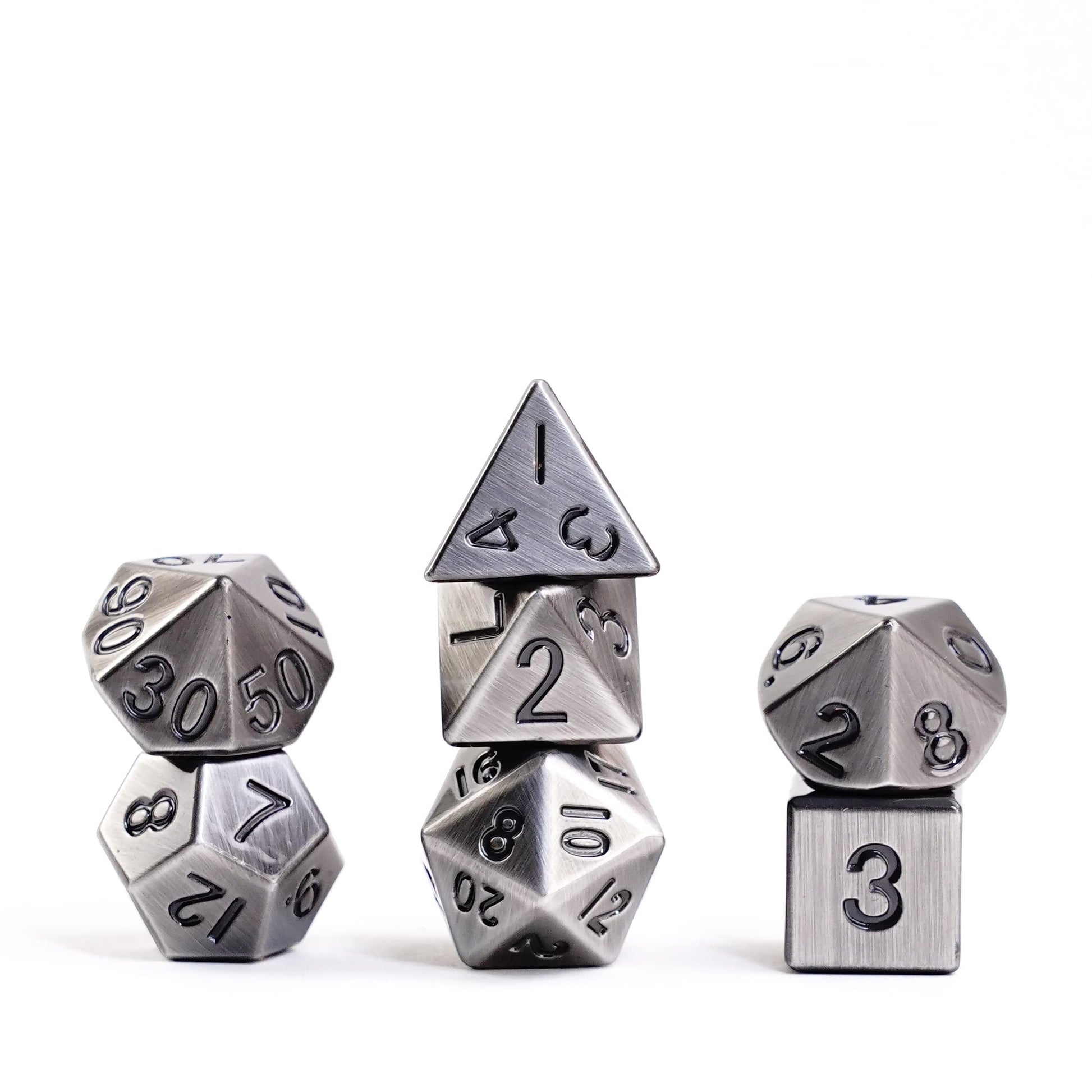 Seven antique silver 16mm metal polyhedral dice in three separate stacks showcase the brushed steel finish and dark engraved numbers on a white background