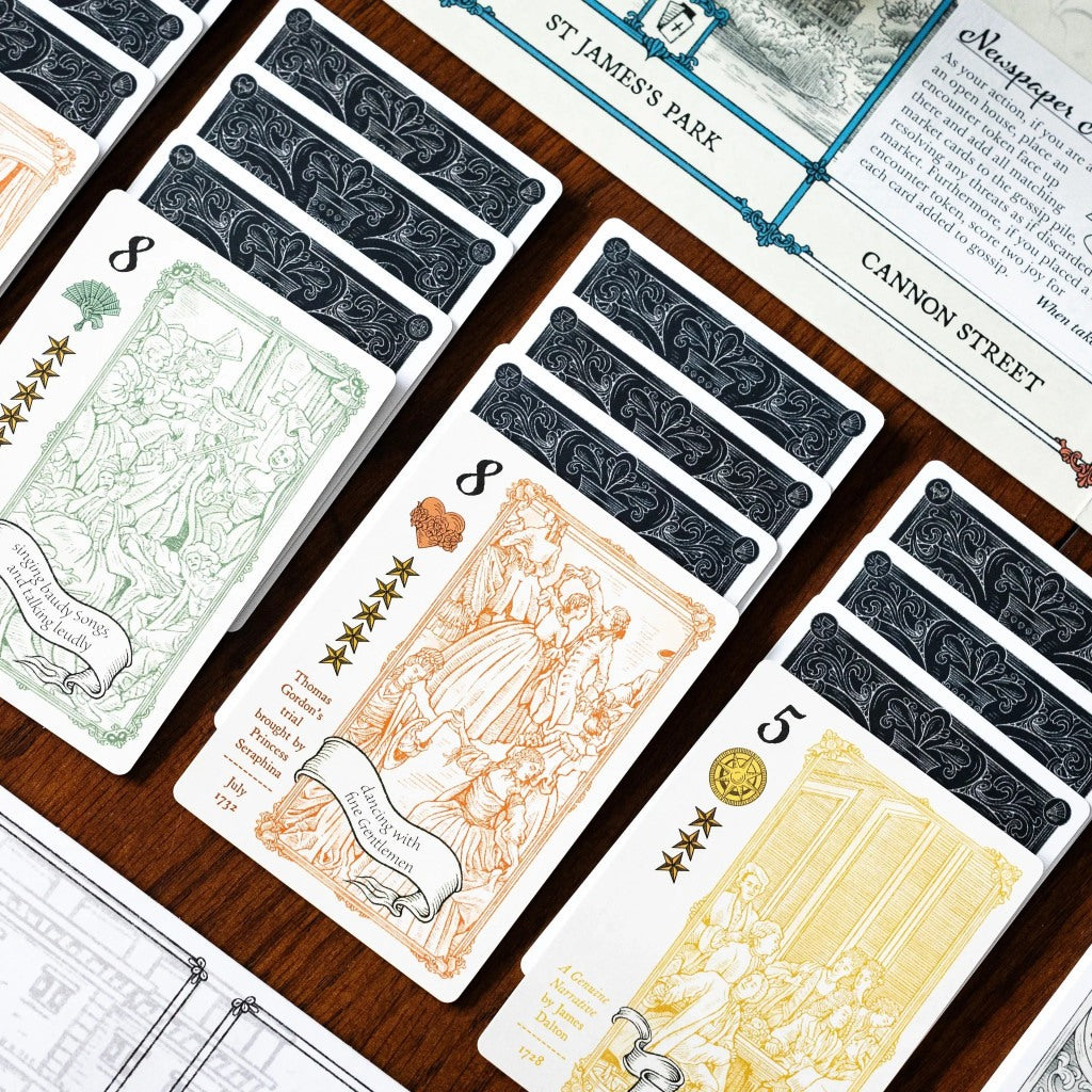 A close-up of custom illustrated game cards featuring vintage orange, green, and yellow line art displayed on a dark wood surface with stacks of ornate dark patterned card backs visible