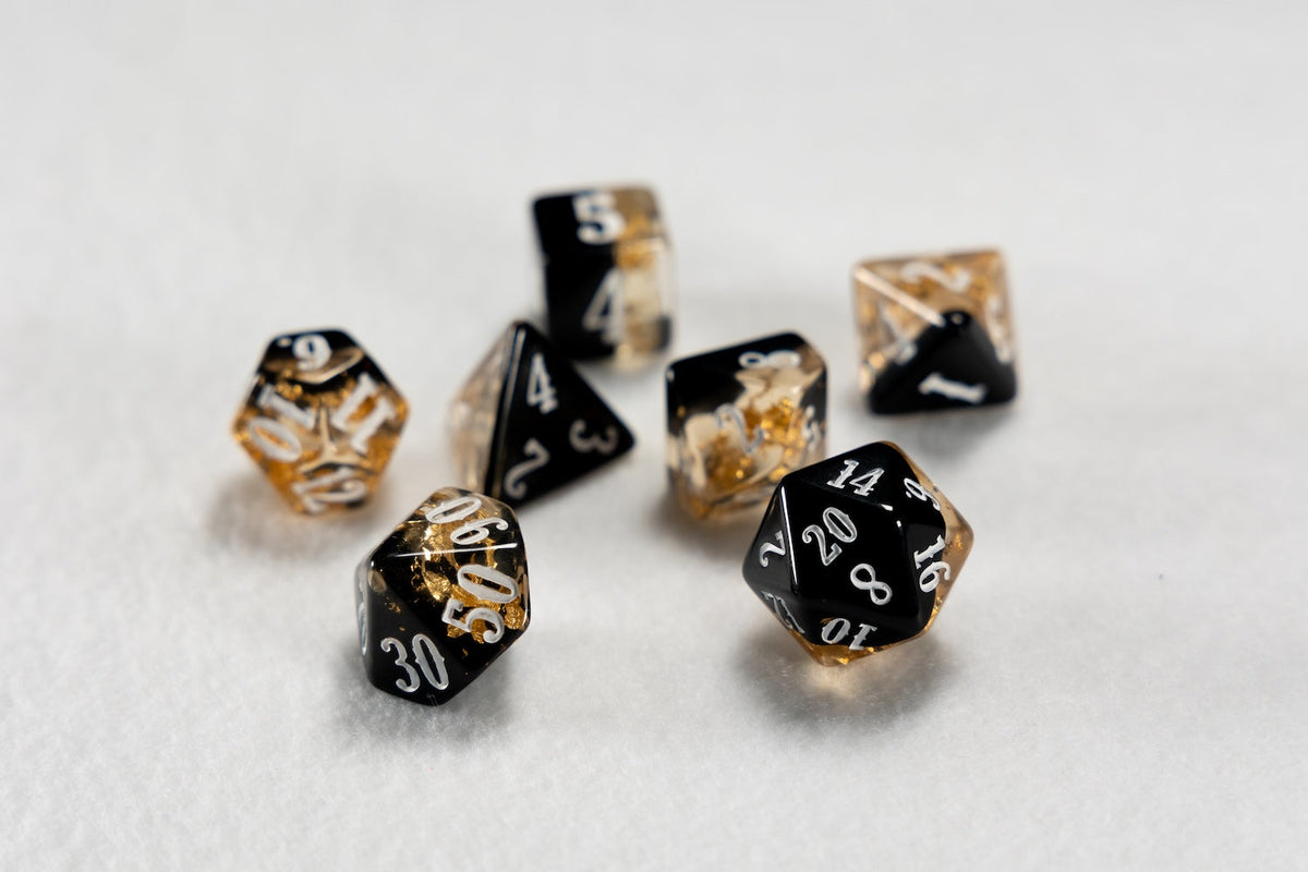 A scattered collection of black and transparent polyhedral dice with gold flakes and white numbers on a light surface