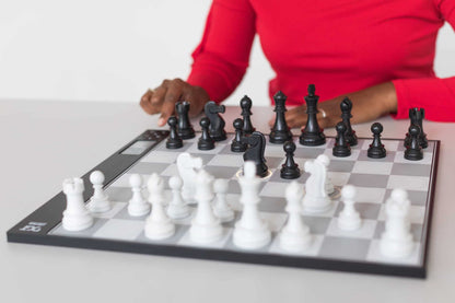 A person in a red shirt plays with a DGT Centaur Chess Computer, a black and gray electronic board with physical white and black pieces, where a black knight's square glows