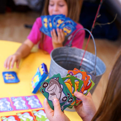 Two players enjoy the Go Fish Card Game, holding hands of colorful cards with cartoon marine life like a smiling shark, set against a yellow table with a metal bucket