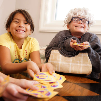 Smiling children are engaged in playing the Continuum Games Old Maid card game, with one child humorously dressed in a wig and glasses