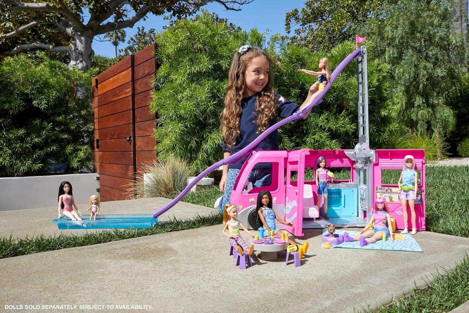 Smiling girl slides a Barbie doll from a pink camper down a purple slide into a pool -  surrounded by other dolls