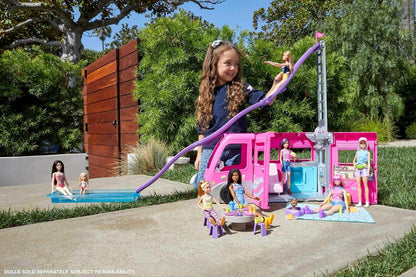 Smiling girl slides a Barbie doll from a pink camper down a purple slide into a pool -  surrounded by other dolls