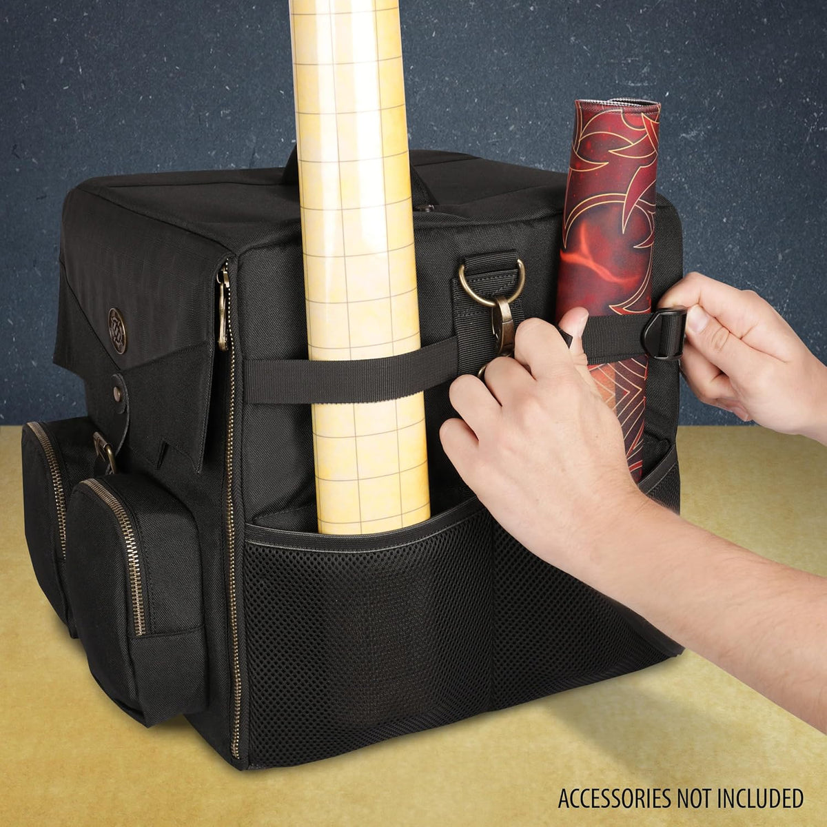 Hands adjust a strap on a black bag, securing a red rolled mat. A yellow grid roll is also in a side pocket.