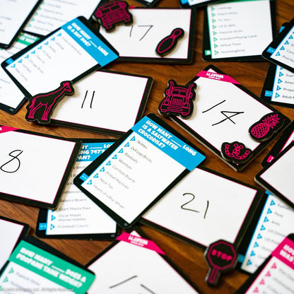 Scattered board game components, including trivia cards, numbered white boards, and neon pink tokens on a wood table