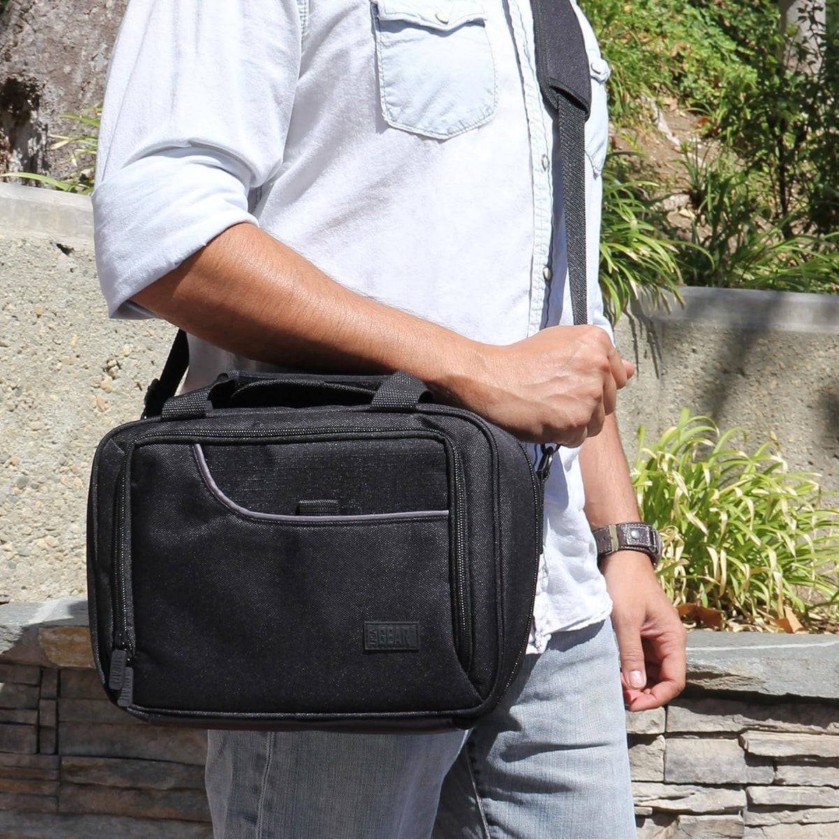 A person in a light denim shirt and jeans carries a black shoulder bag with a gray trim outdoors