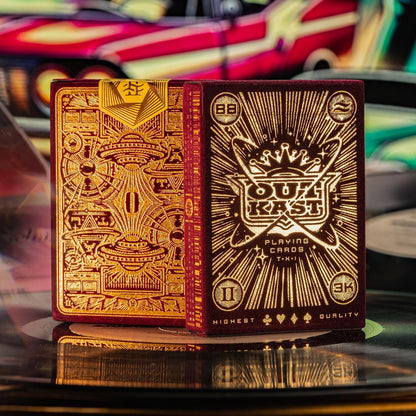 Two red velvet playing card tuck boxes with detailed gold foil stamping, featuring Outkast branding and UFO imagery, sitting on a record