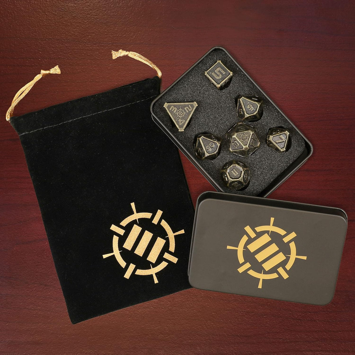 Dark metal polyhedral dice set in an open tin, with a black velvet bag and closed metal tin. All have a gold logo