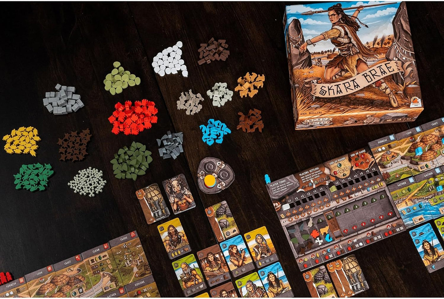 Skara Brae board game display, including the box, resource tokens, character cards, and modular game boards on a dark wooden table
