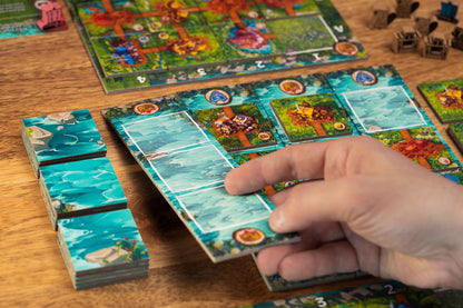 A hand places a blue water tile onto the Timber Town board game, which has colorful land tiles and stacks of water tiles nearby