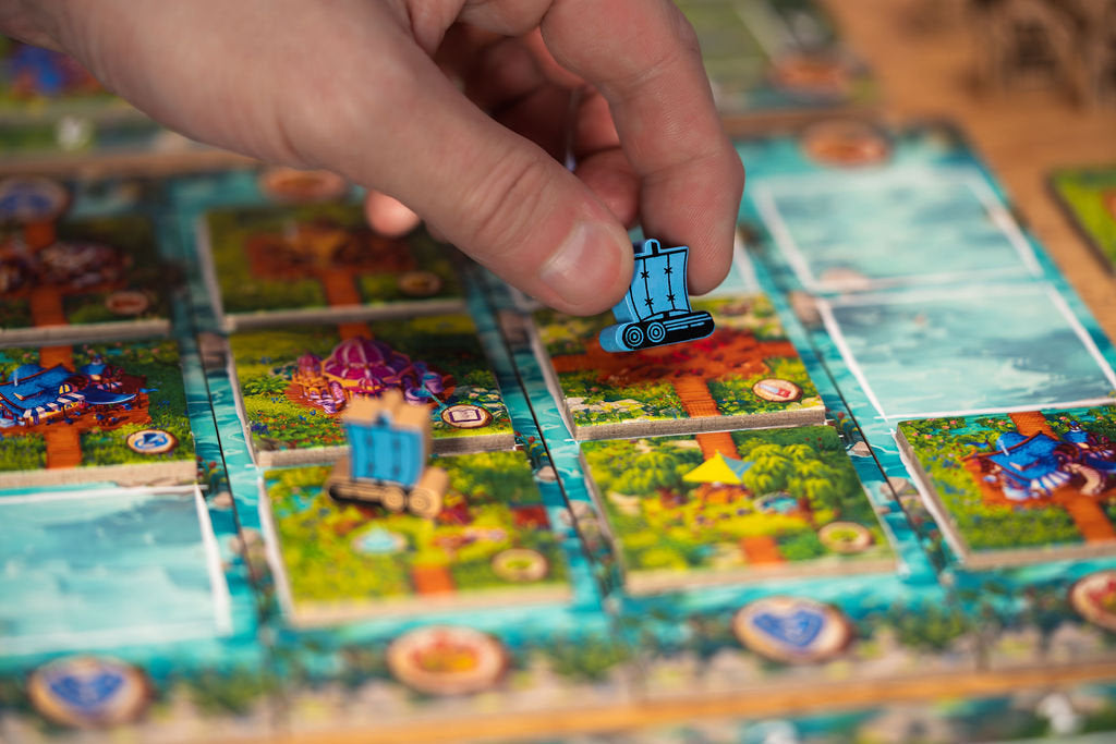 A hand places a blue wooden ship game piece onto a colorful Timber Town board game featuring island and water tiles