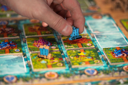 A hand places a blue wooden ship game piece onto a colorful Timber Town board game featuring island and water tiles