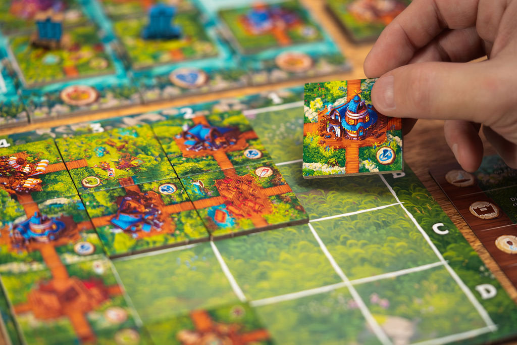 A hand places a Timber Town game tile featuring a blue house onto a green grid board