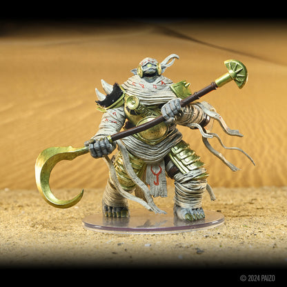 Large mummified creature in gold armor holds a golden two-ended polearm, standing on a sandy desert base