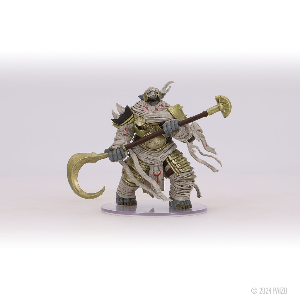 A hulking, bandaged creature miniature with gold armor and a horned head, wielding a large staff with a curved golden blade