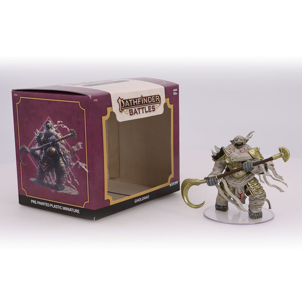 A Pathfinder Gholdako miniature, bandaged and armored, holding a golden staff, next to its open box