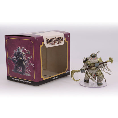 A Pathfinder Gholdako miniature, bandaged and armored, holding a golden staff, next to its open box