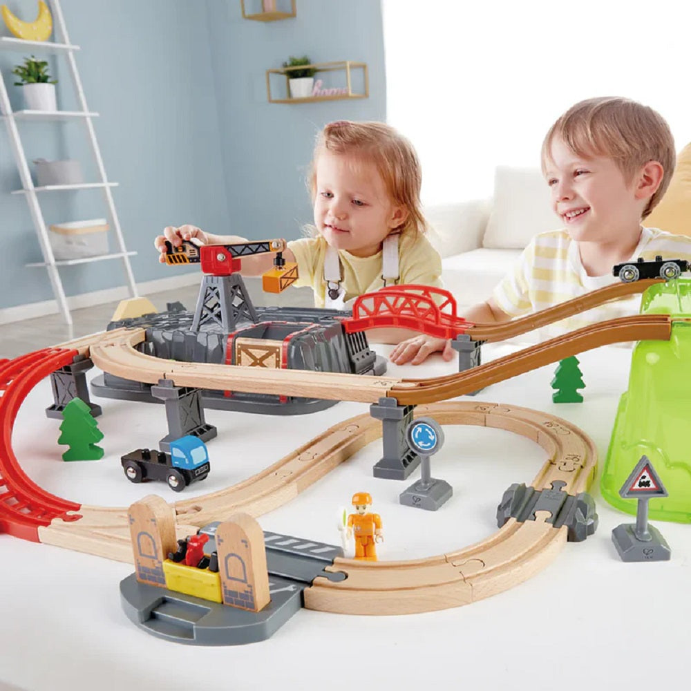 Two happy children playing with a large construction-themed wooden toy train track and crane set