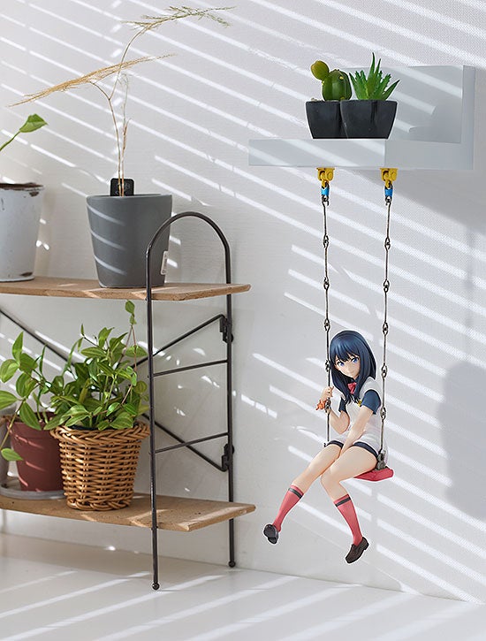 Anime schoolgirl figure on a tiny red swing hanging from a white shelf with miniature potted plants