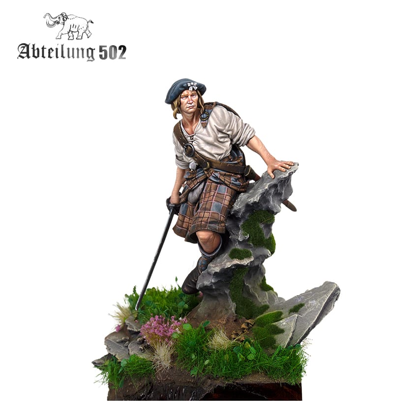 A detailed miniature figure depicts a Highlander in kilt and beret, holding a sword on a rocky, mossy base with flowers