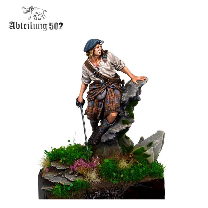 A detailed miniature Highlander figure wearing a kilt and beret, holding a sword, leaning on a mossy rock base with grass and purple flowers
