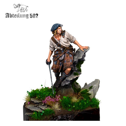 A detailed miniature Highlander figure wearing a kilt and beret, holding a sword, leaning on a mossy rock base with grass and purple flowers