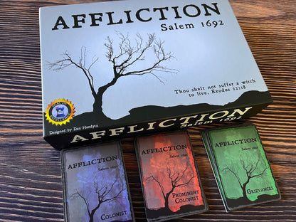 Overhead view of "Affliction Salem 1692" board game box with bare tree art, and three matching card decks on a wooden table