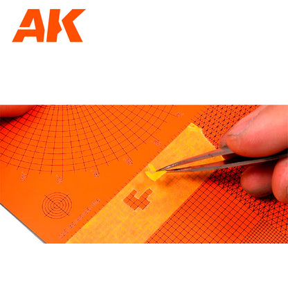 AK logo. A hand uses tweezers to precisely place a small yellow masking tape detail onto an orange grid crafting mat